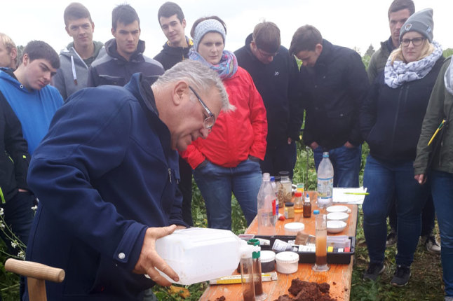 Mann steht vor Biertisch mit Versuchsgegenständen, hinter ihm im Halbrund junge Leute